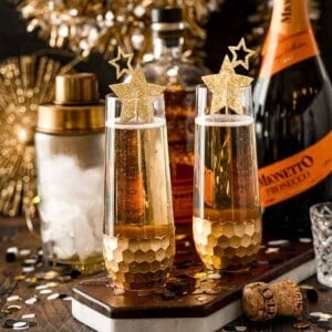 Two stemless champagne classes with a shaker, ice, bourbon, and prosecco in the background. Drinks are garnished with gold stars.