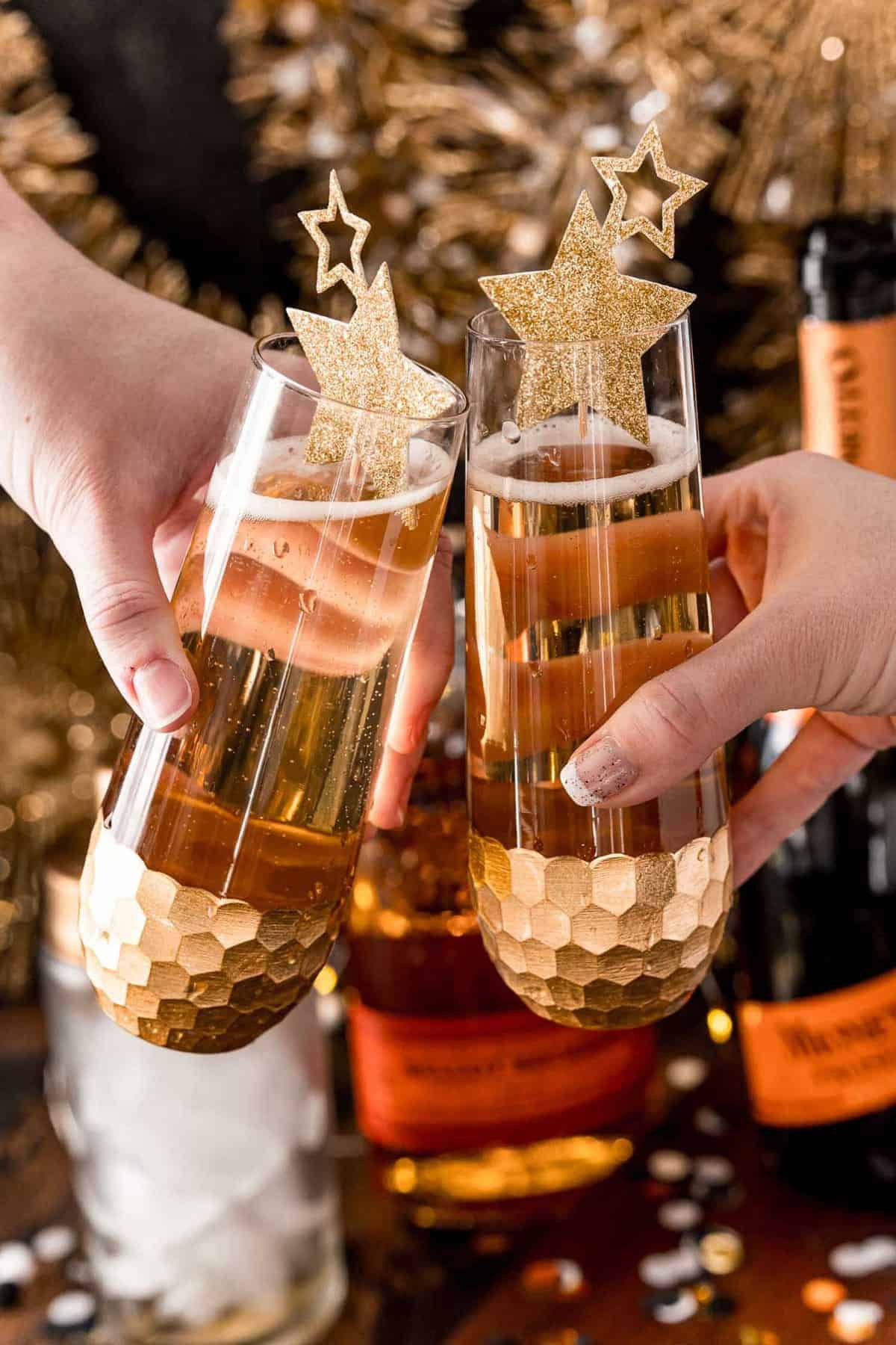 Two hands holding stemless champagne flutes filled with a light brown drink.