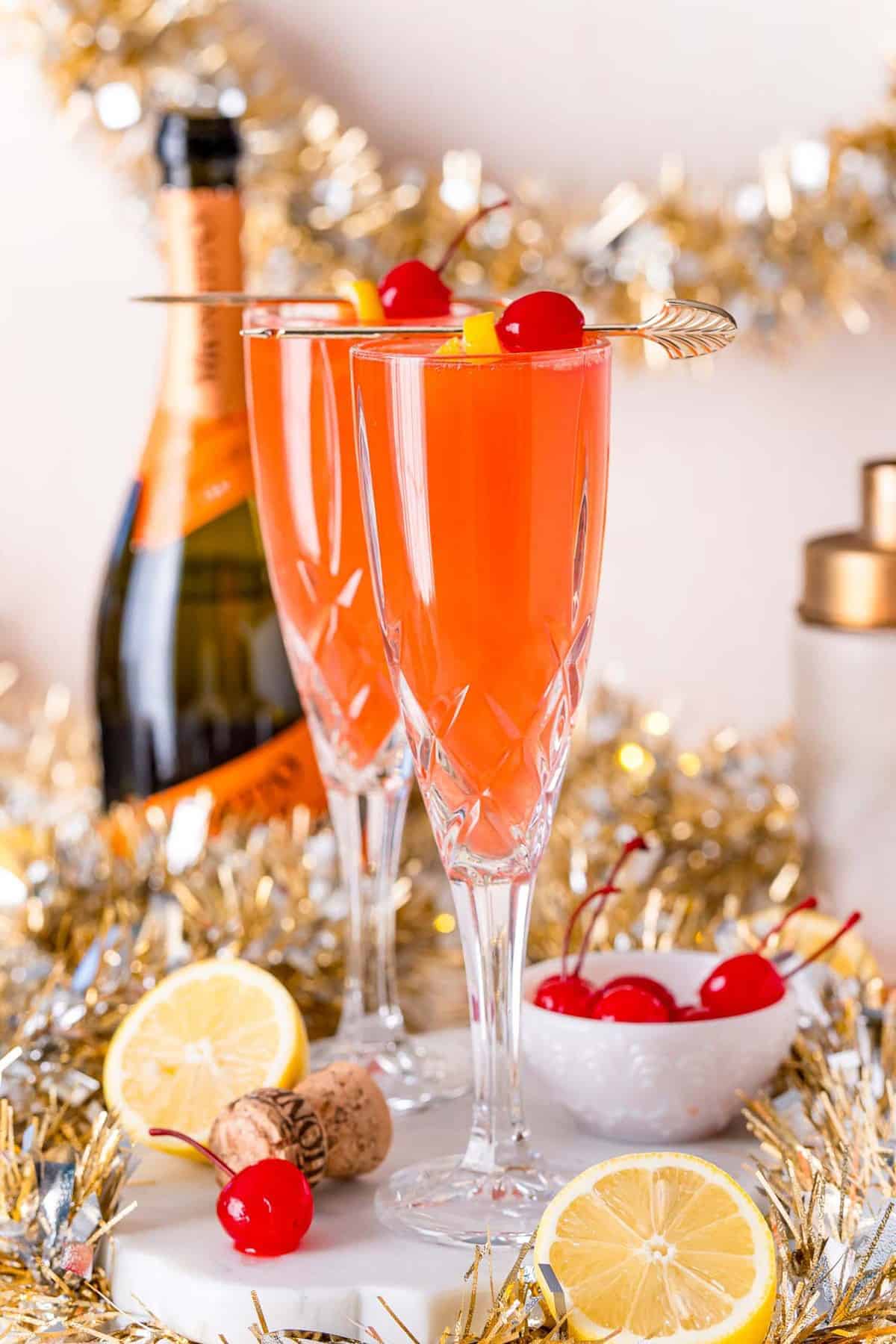 Two champagne flutes with a light red drink garnished with cherries and lemon.