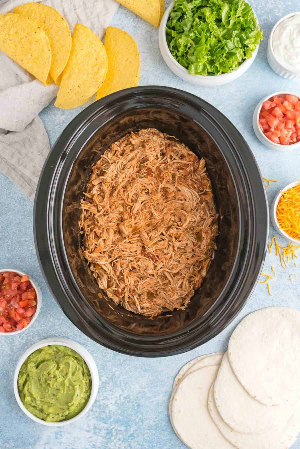 Taco chicken in a slow cooker, surrounded with toppings.