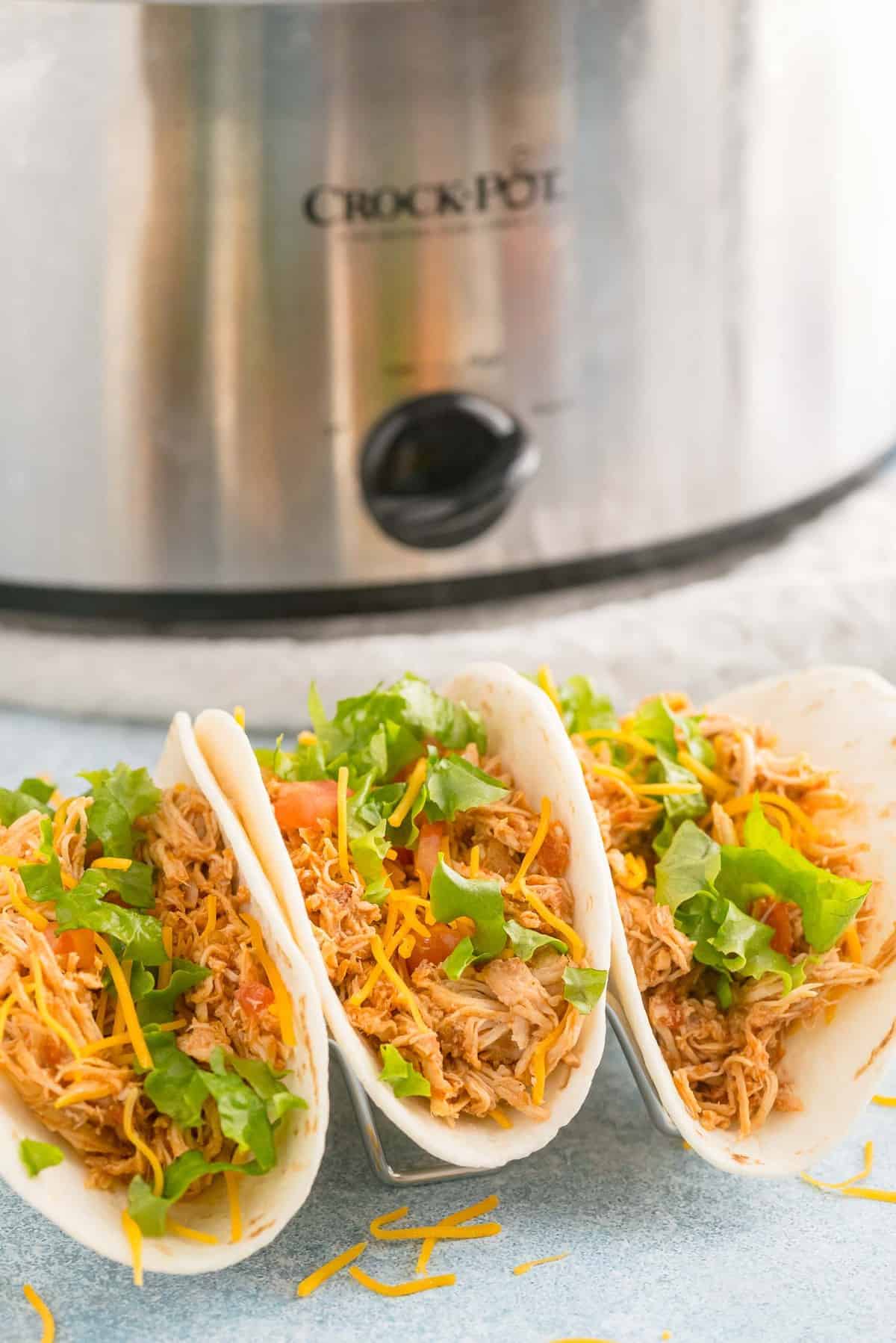 Three soft chicken tacos in front of a crockpot.