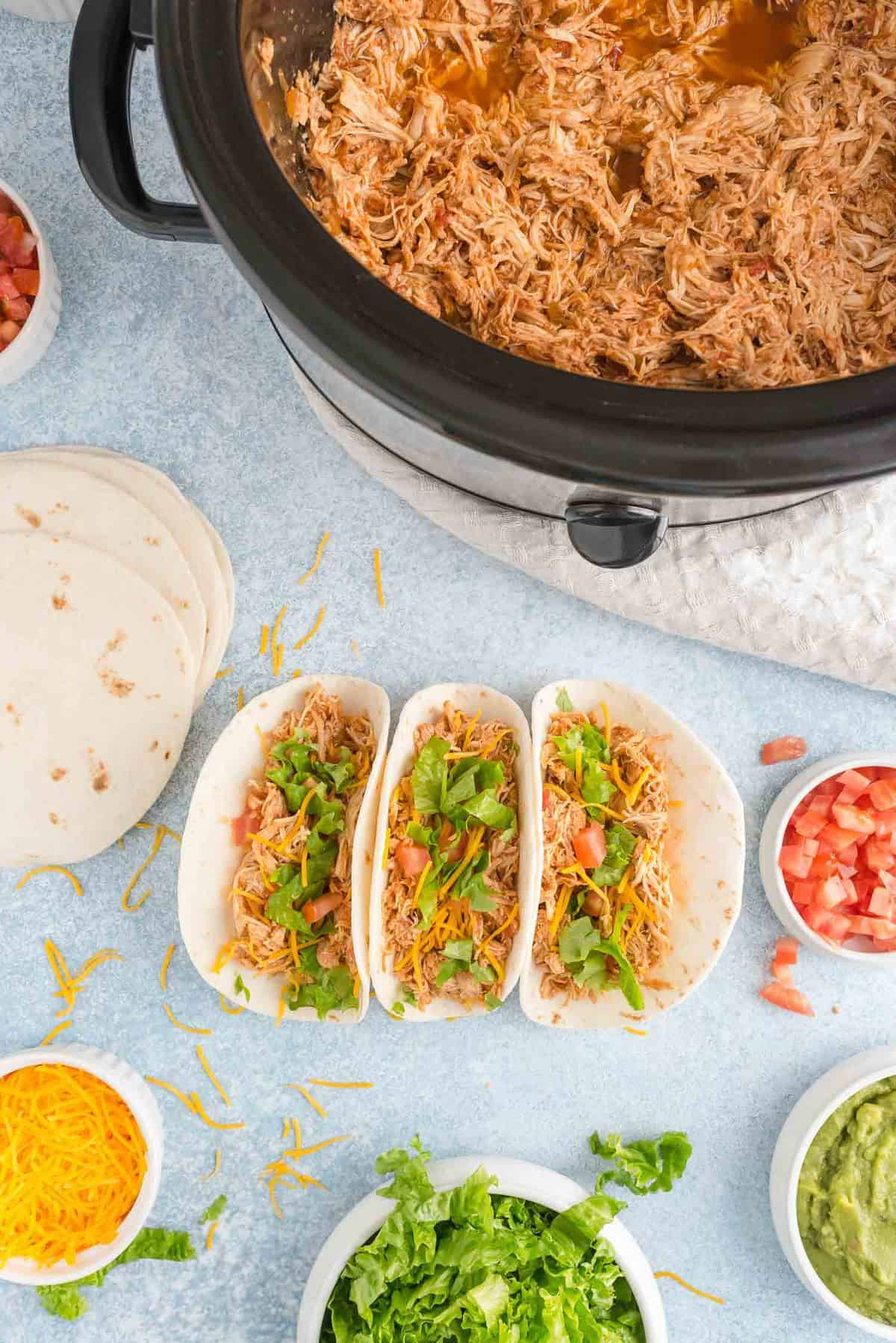 Overhead view of crockpot taco chicken, some in slow cooker and some composed in tacos.