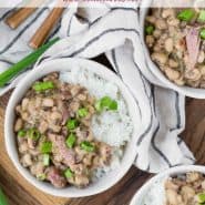 Beans, rice, and ham topped with green onions in small white bowls. Text overlay reads "hoppin' john (black eyed peas & rice), rachelcooks.com"