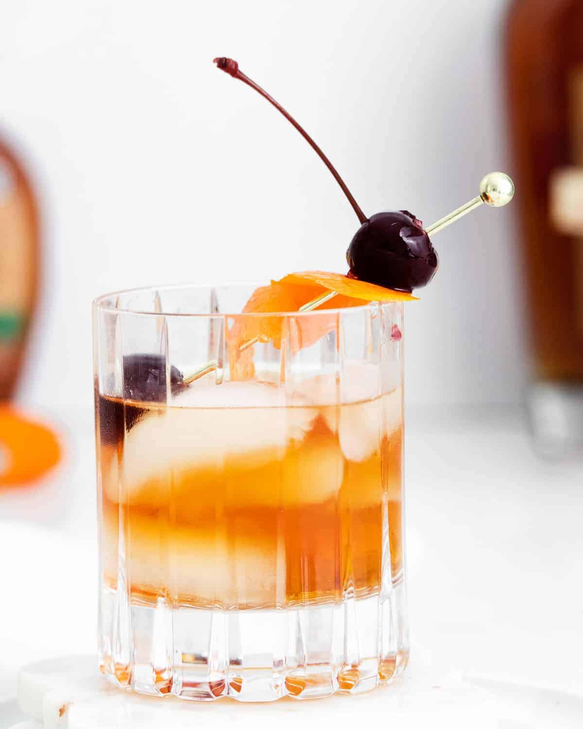 Short cocktail glass with ice, orange peel, cherry, and whiskey.