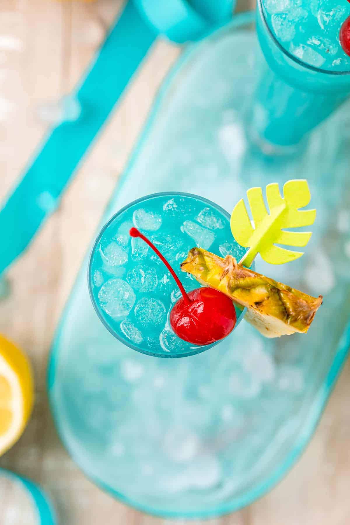 Overhead view of bright blue drink with a pineapple wedge and a cherry.