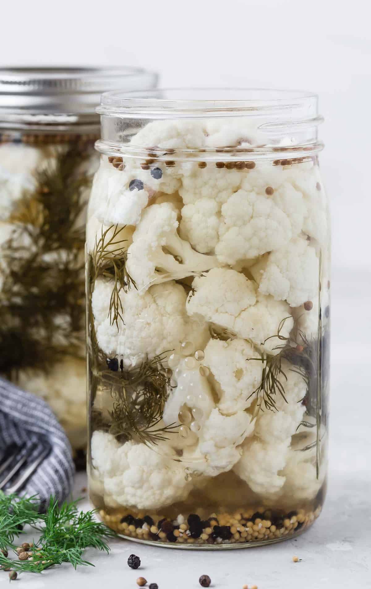Jar of pickled cauliflower with dill and peppercorns, another jar in background.