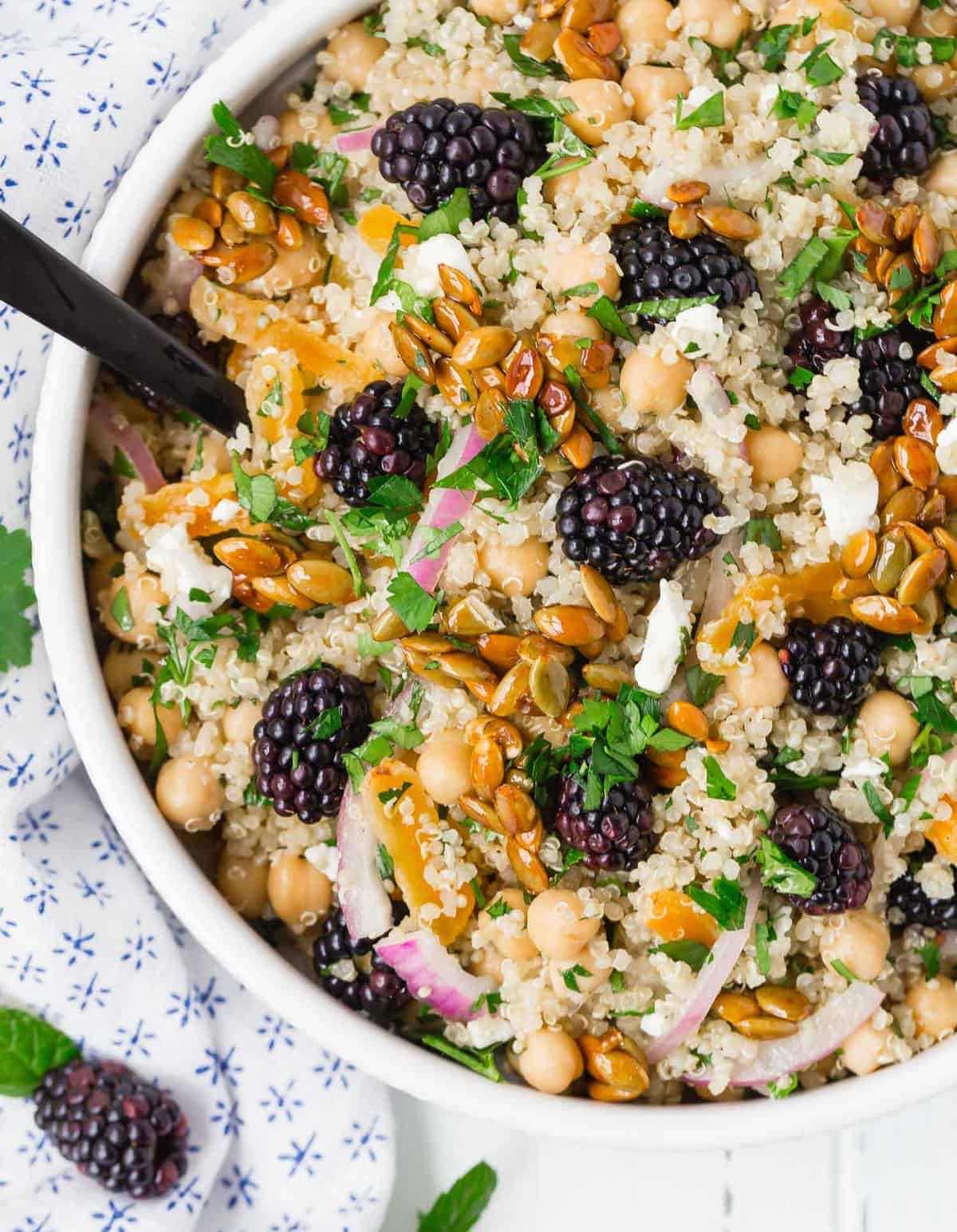 Close up of a salad in a white bowl comprised of quinoa, chickpeas, feta cheese, red onion, blackberries and more.