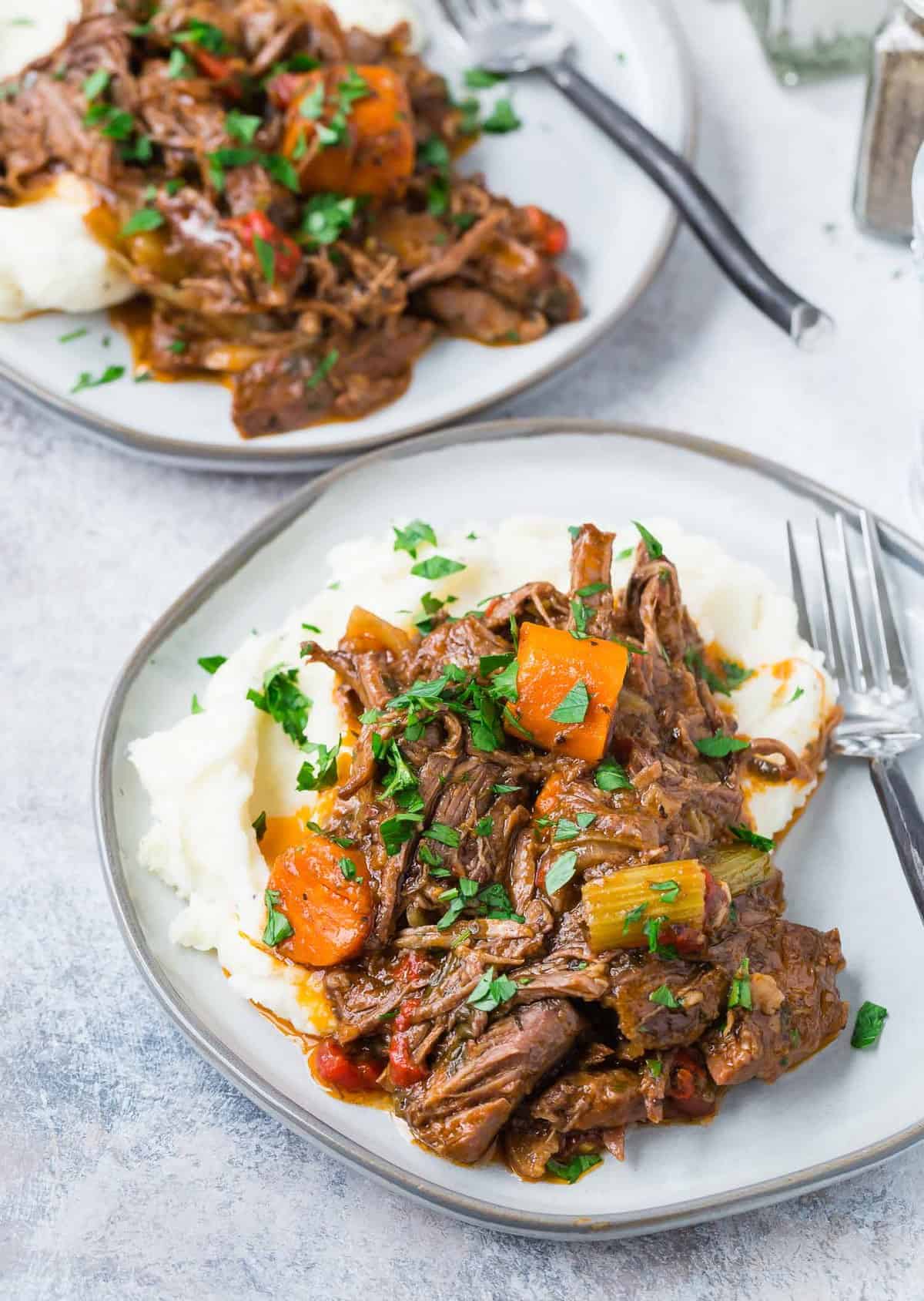 Pot roast served over potatoes on two gray plates.