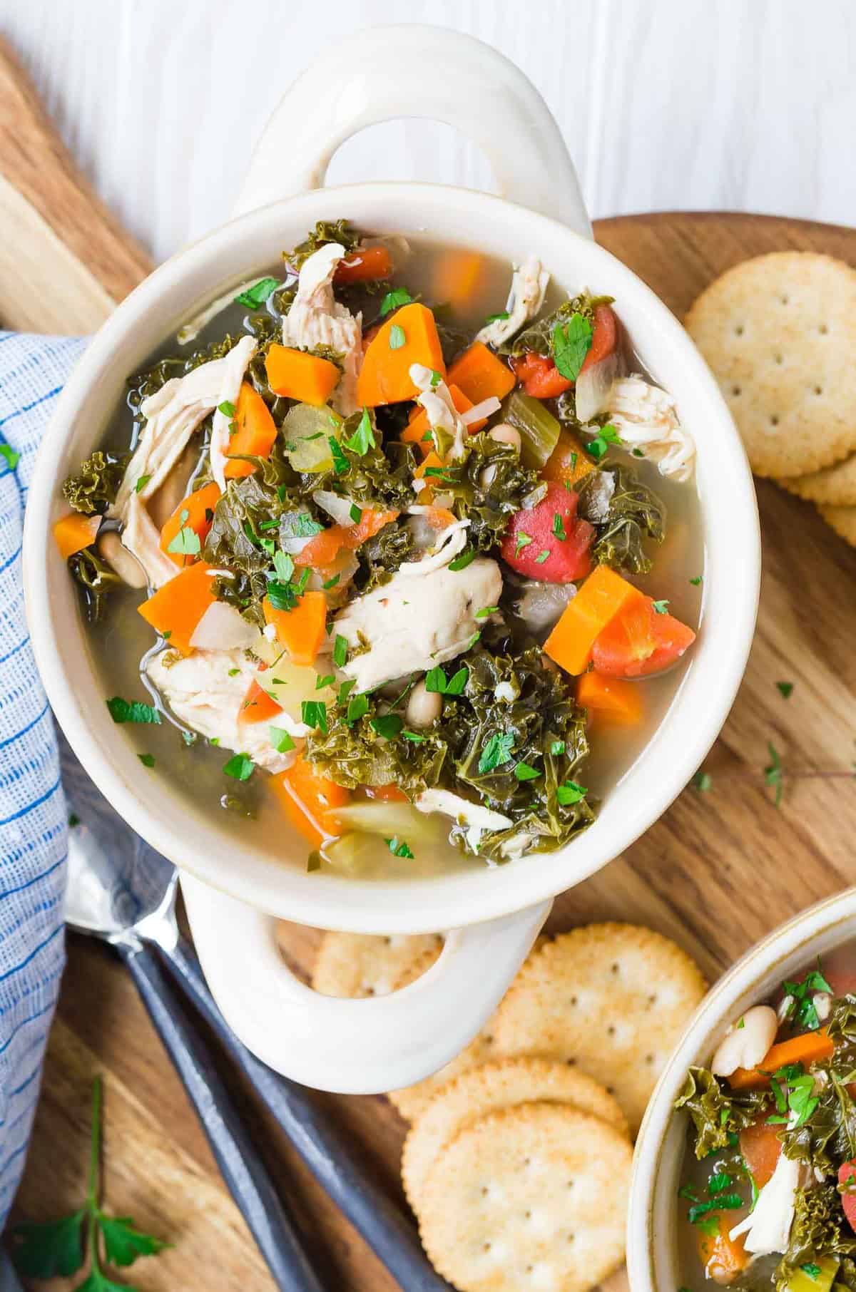 Overhead view of a bowl of soup in a two-handled bowl. Soup includes kale, chicken, carrots.