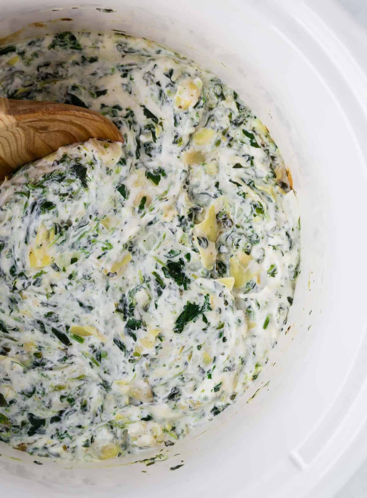 Overhead view of spinach dip with artichoke hearts in a white slow cooker with a wooden spoon.