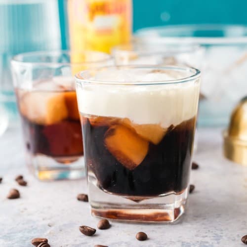 A white Russian cocktail in a short glass, with coffee beans scattered around it.