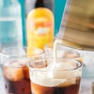 A short clear glass with a dark brown liquid, and heavy cream being poured in. Text overlay reads "how to make a white russian, rachelcooks.com"