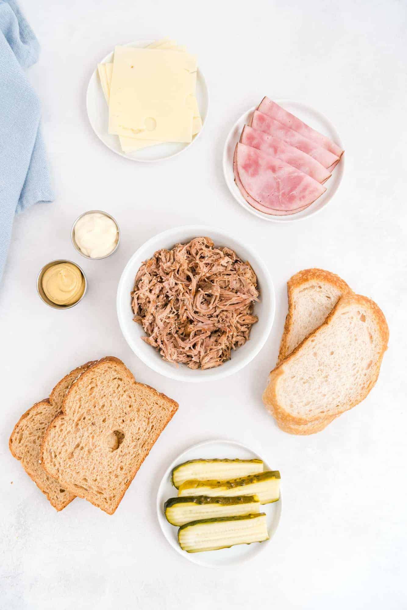 Overhead view of ingredients: pork, ham, pickles, bread, mustard, mayo, cheese.