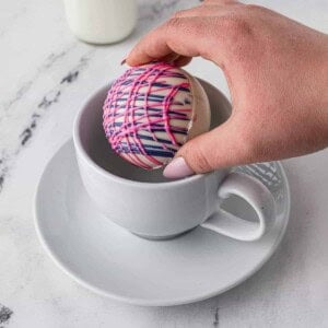 A hot cocoa bomb being added to a white mug on a saucer.