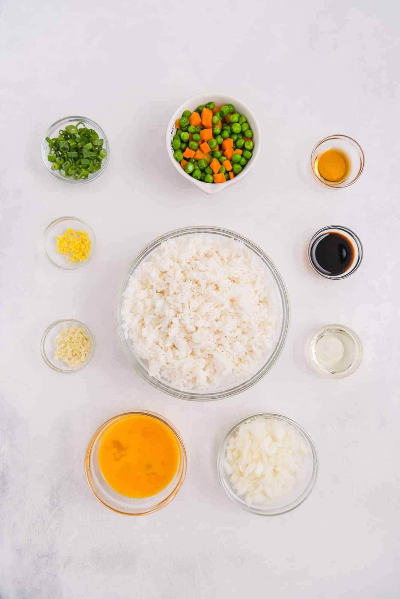 Overhead view of ingredients in small bowls: rice, eggs, vegetables, soy sauce, green onions, oil.