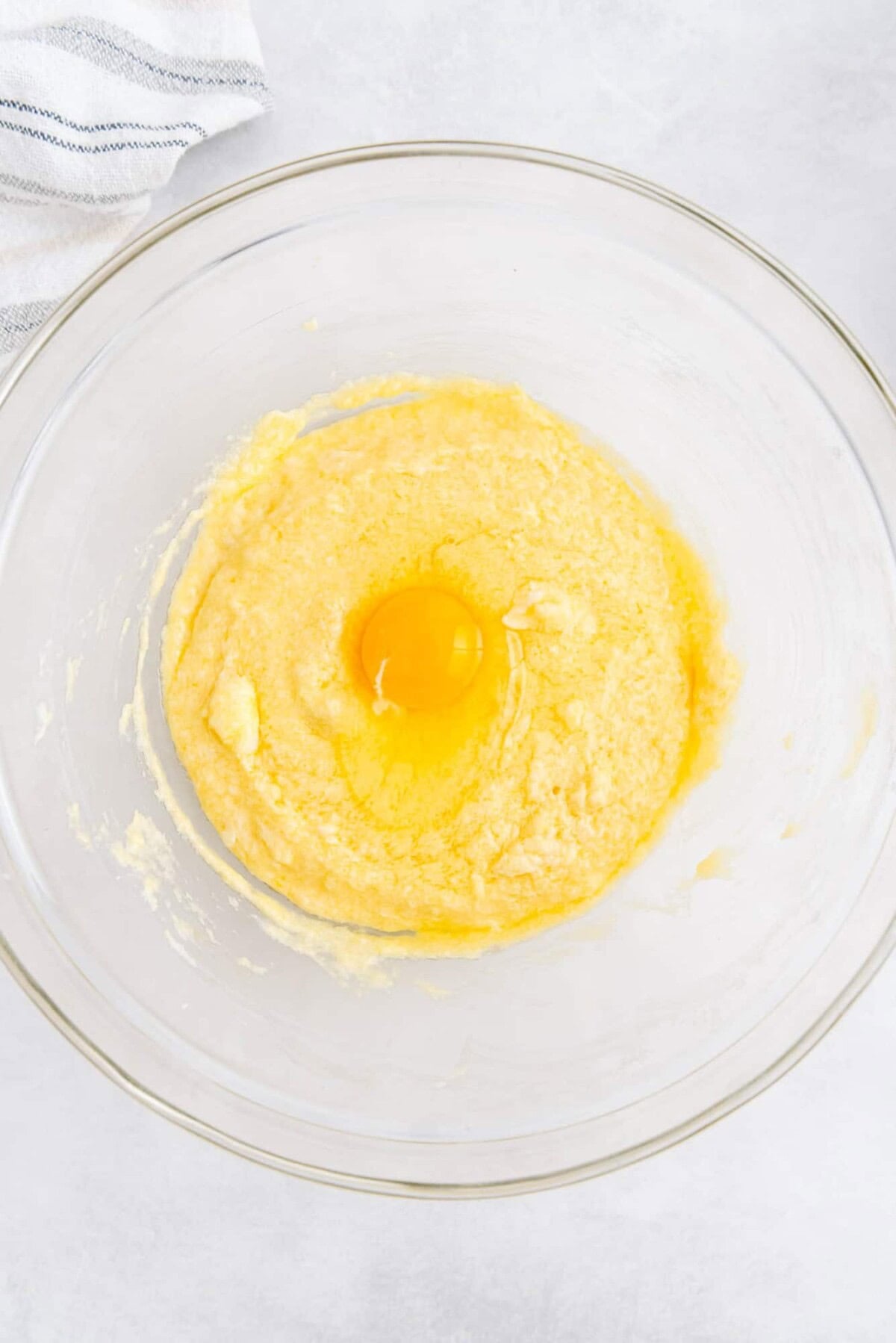 A clear glass bowl contains a yellow batter mixture with a raw egg cracked on top, ready to be mixed.