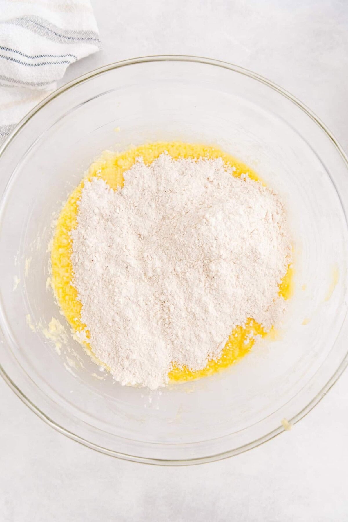 A glass bowl containing a mixture of flour and yellow wet ingredients sits on a light surface with a striped towel partly visible in the corner.