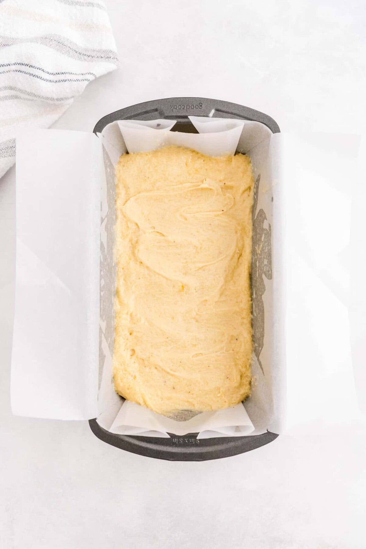 A metal loaf pan lined with parchment paper is filled with unbaked yellow batter, sitting on a light-colored surface next to a striped towel.