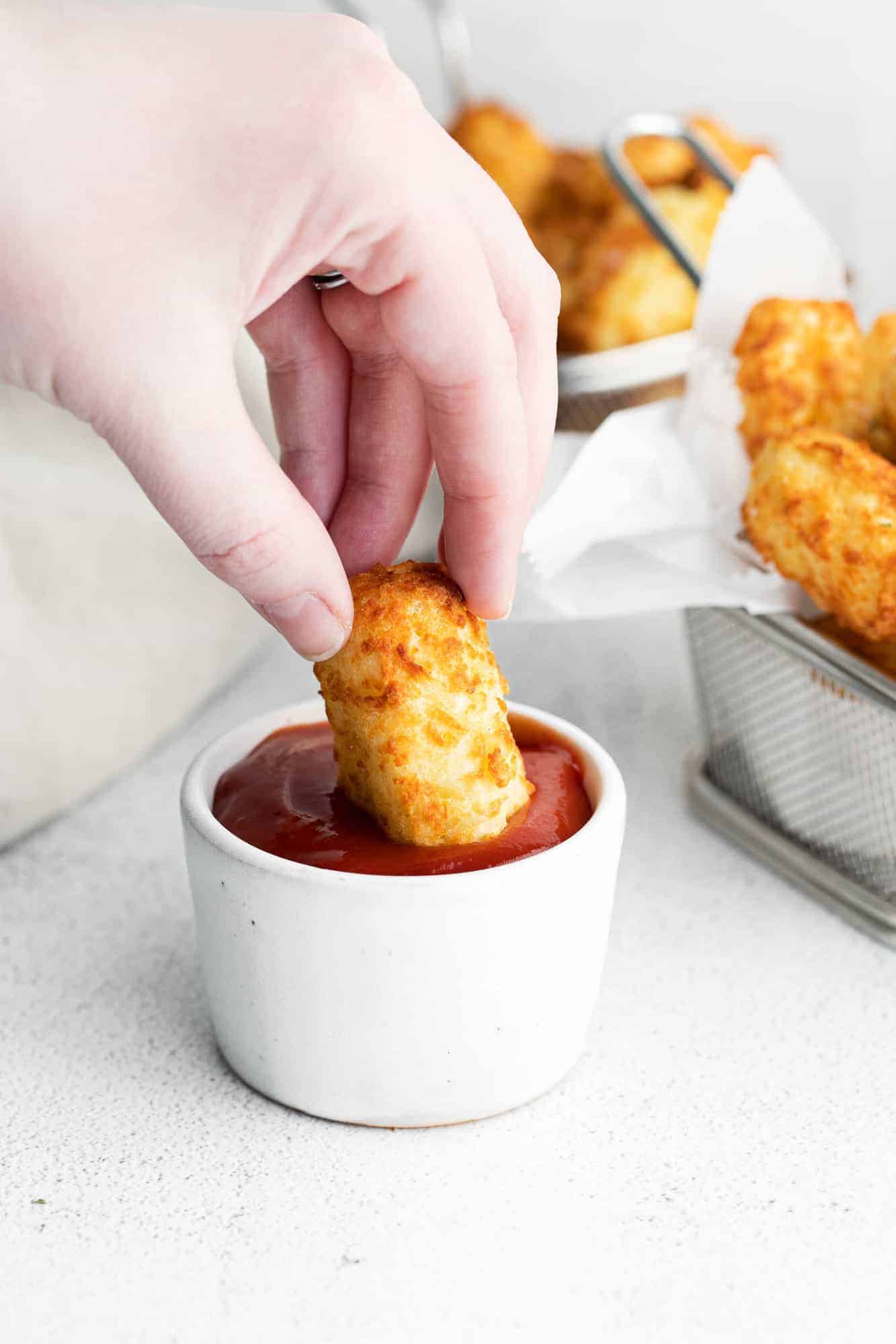 Tater tot being dipped into a small white dish of ketchup.