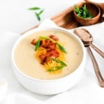Cauliflower soup in a white bowl, topped with green onions, cheddar, and bacon. Spoons also visible.