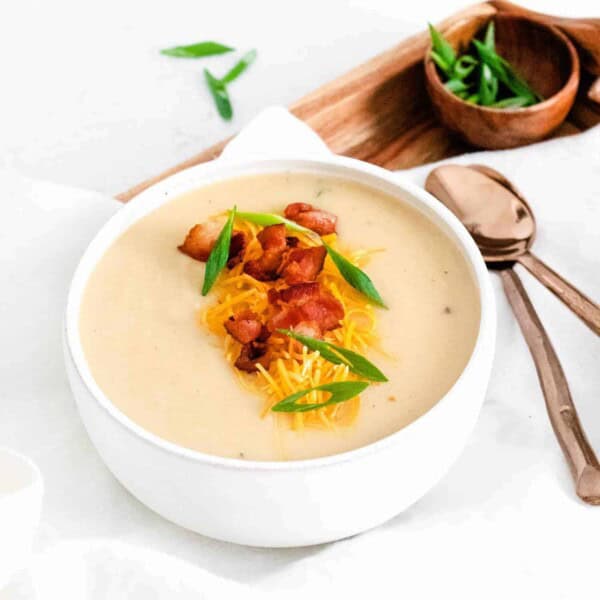 Cauliflower soup in a white bowl, topped with green onions, cheddar, and bacon. Spoons also visible.