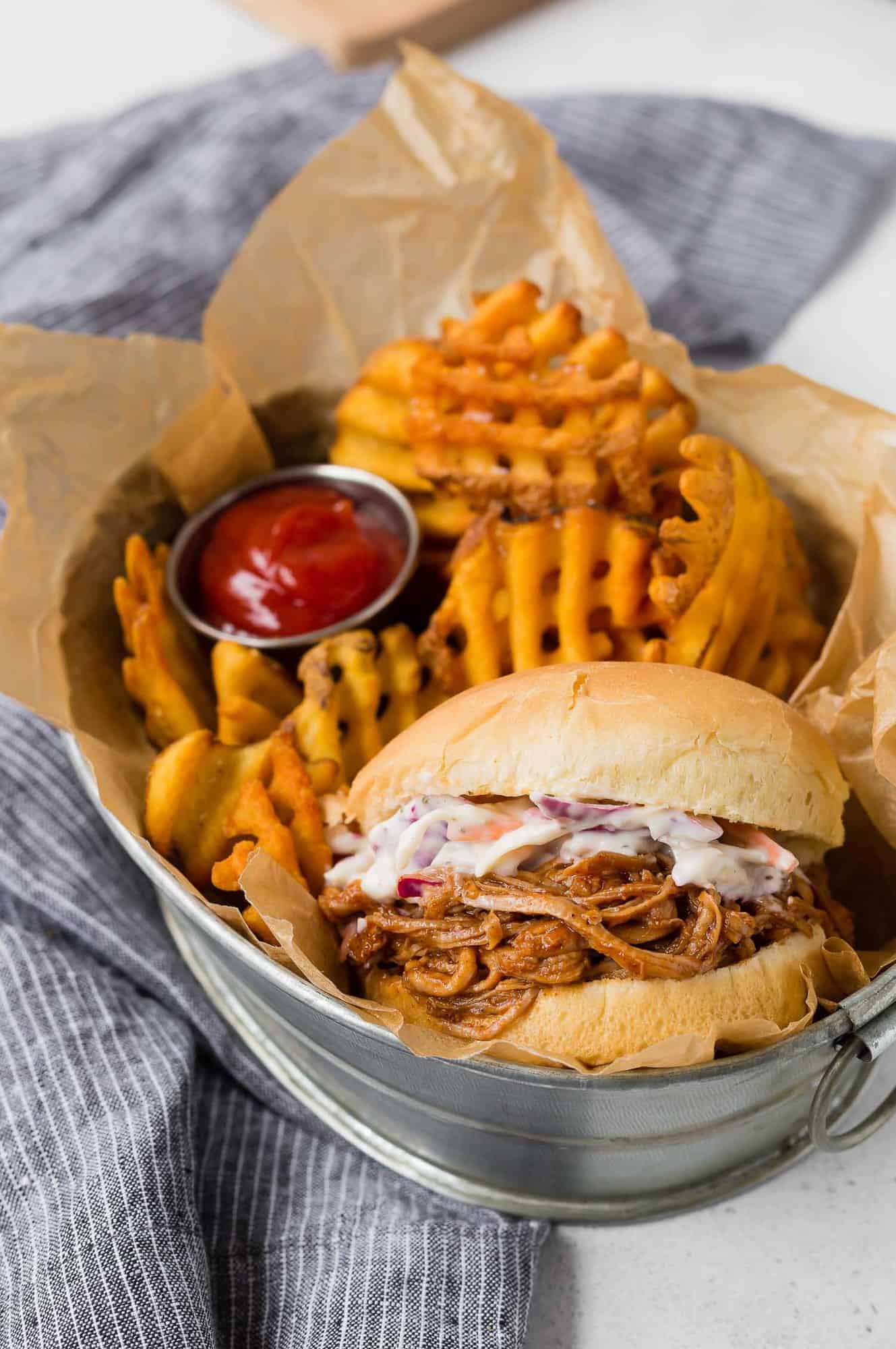 Pork sandwich, waffle fries, and ketchup in a metal basket.