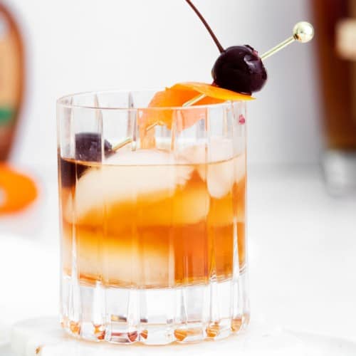 An old fashioned cocktail on ice with an orange peel and cherries.