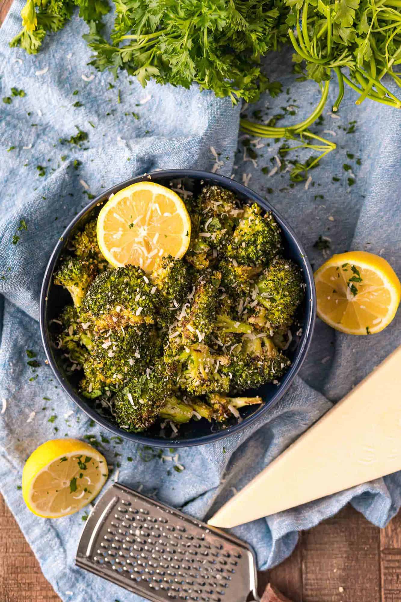 Roasted broccoli in black bowl, surrounded by lemons, cheese, parsley.