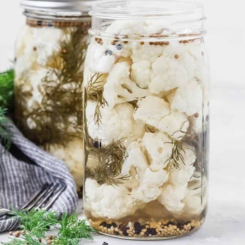 Two jars of cauliflower florets in pickling liquid with whole spices and dill.