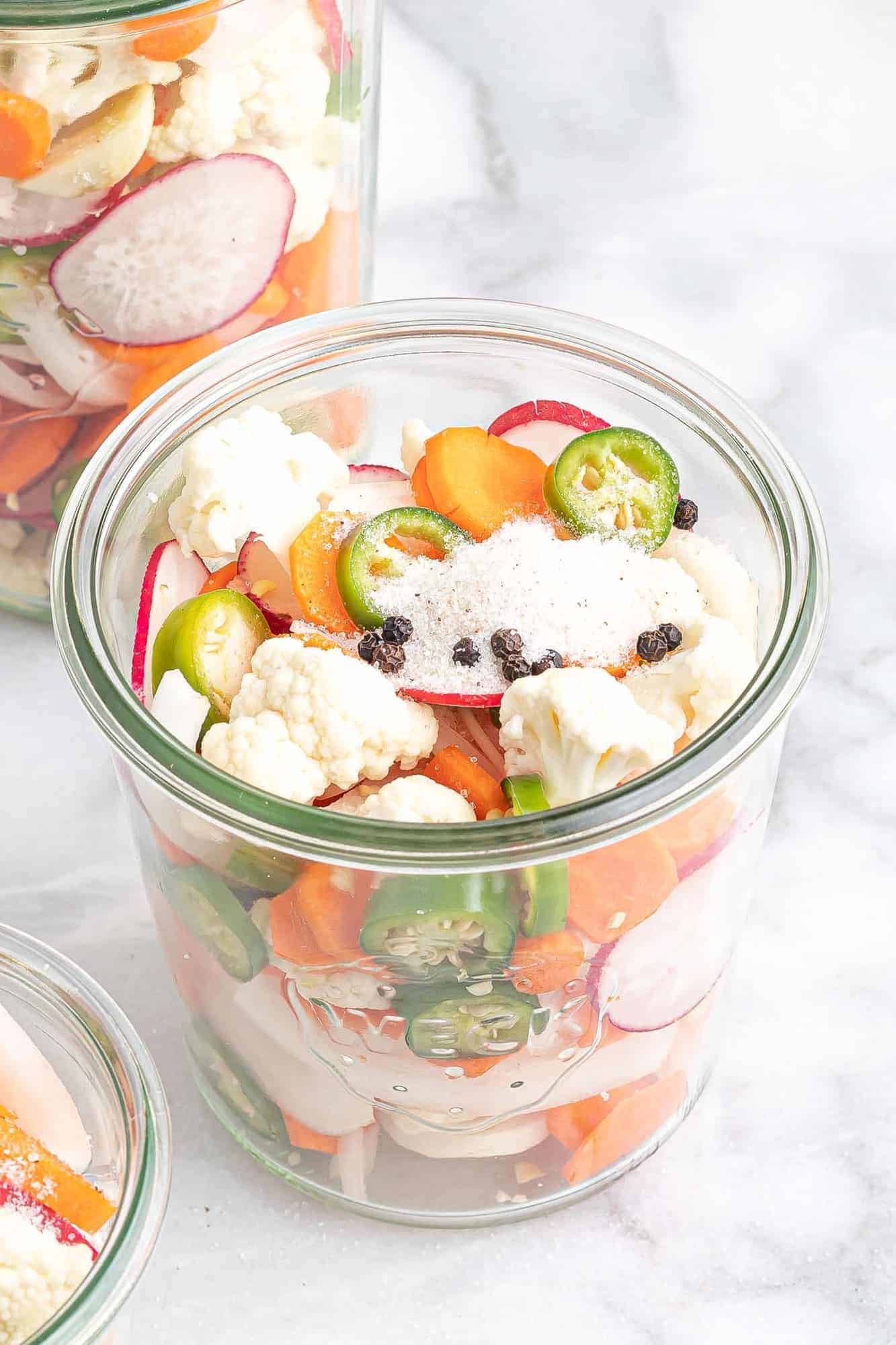 Cut vegetables, topped with sugar, salt, and peppercorns, in a glass jar.