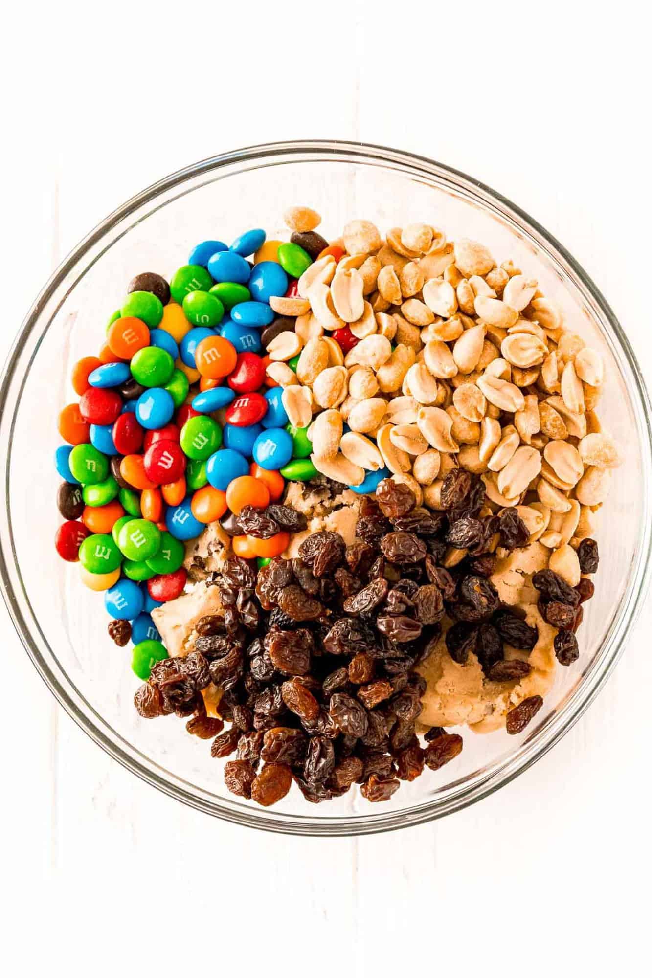 Unmixed cookie dough, raisins, m&m's, and peanuts in a glass bowl.