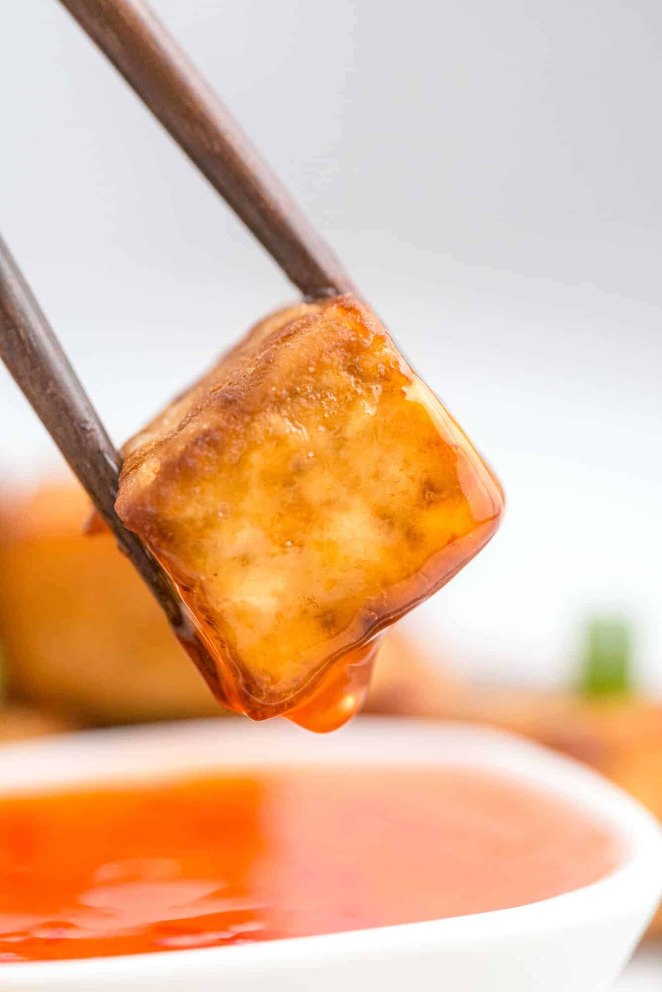 Tofu cube between chopsticks, with a drip of sweet chili sauce dripping off it.