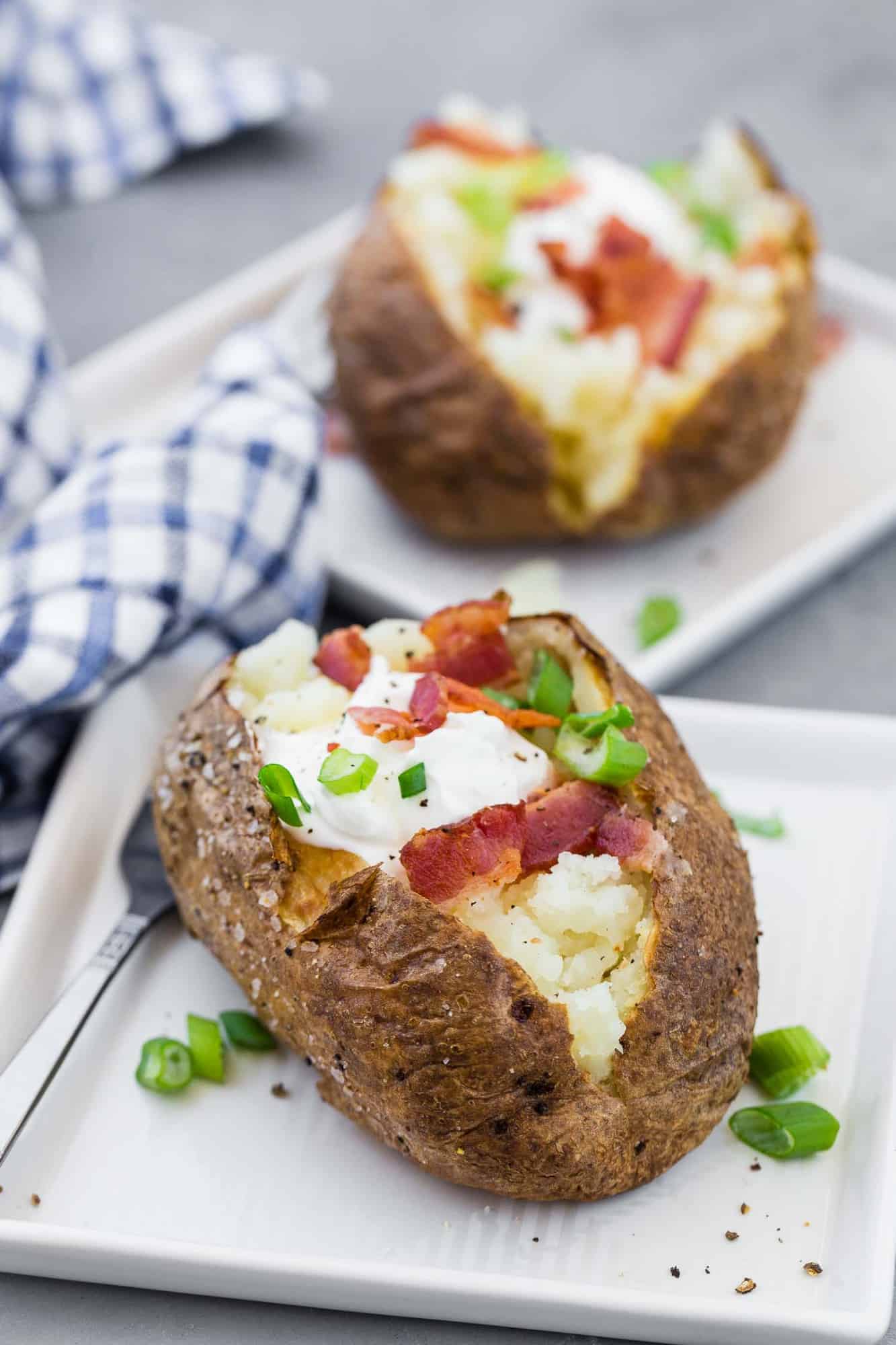 Baked potatoes on white plates, topped with sour cream, green onions, and bacon.