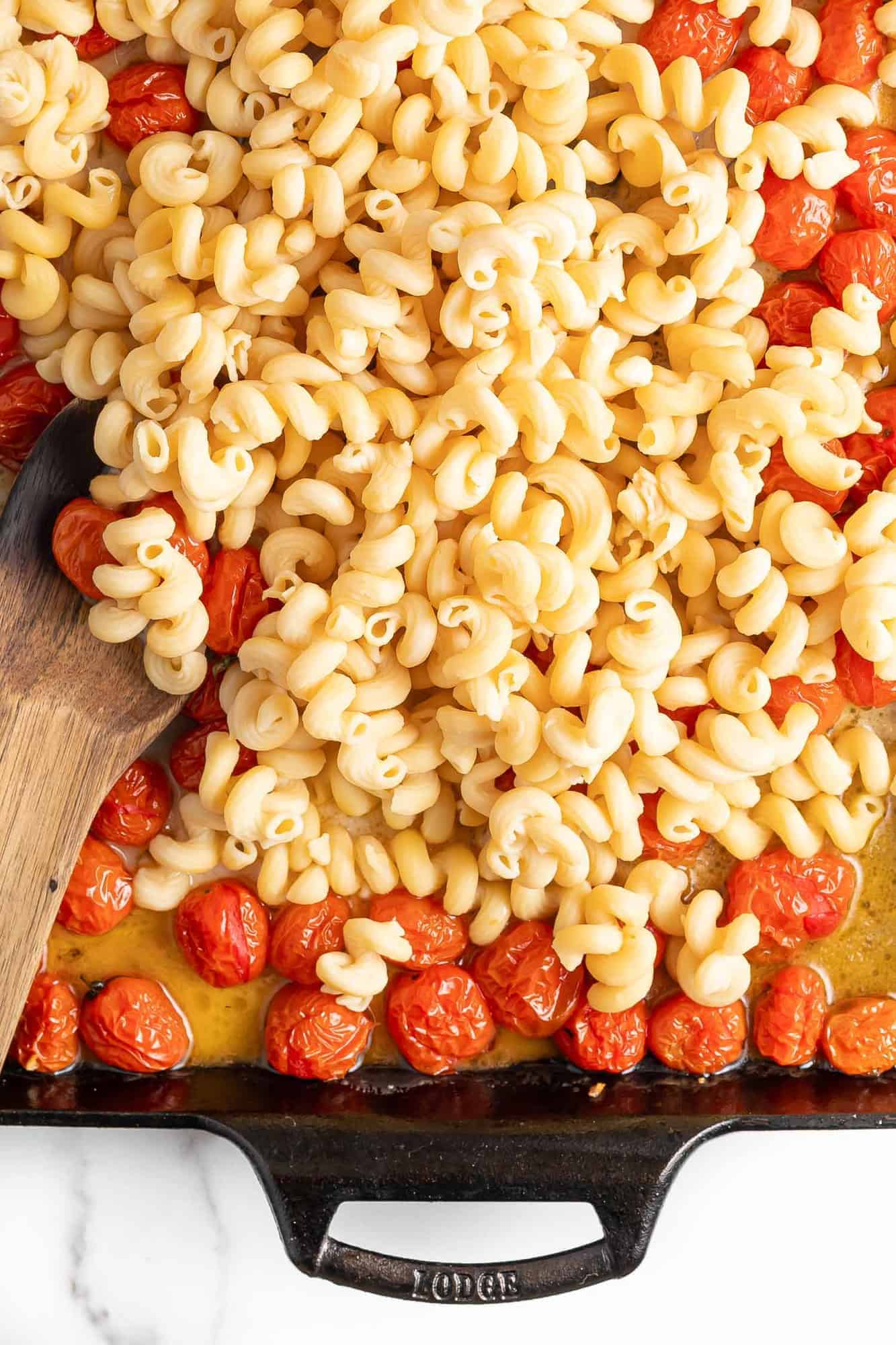 Pasta being stirred into roasted tomatoes and feta in a baking dish.