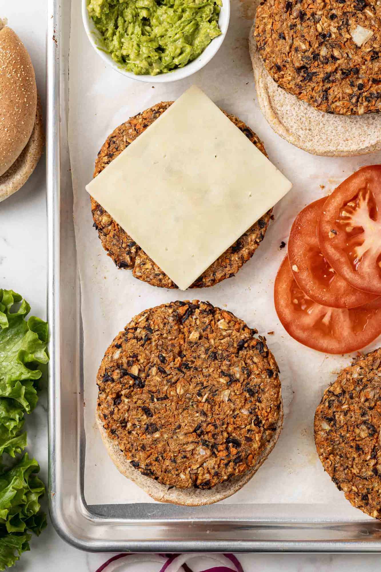 Bean burgers on a baking sheet, one with a slice of cheese on it. Toppings also visible.