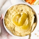 Hummus in a white bowl with a spoon, topped with olive oil.