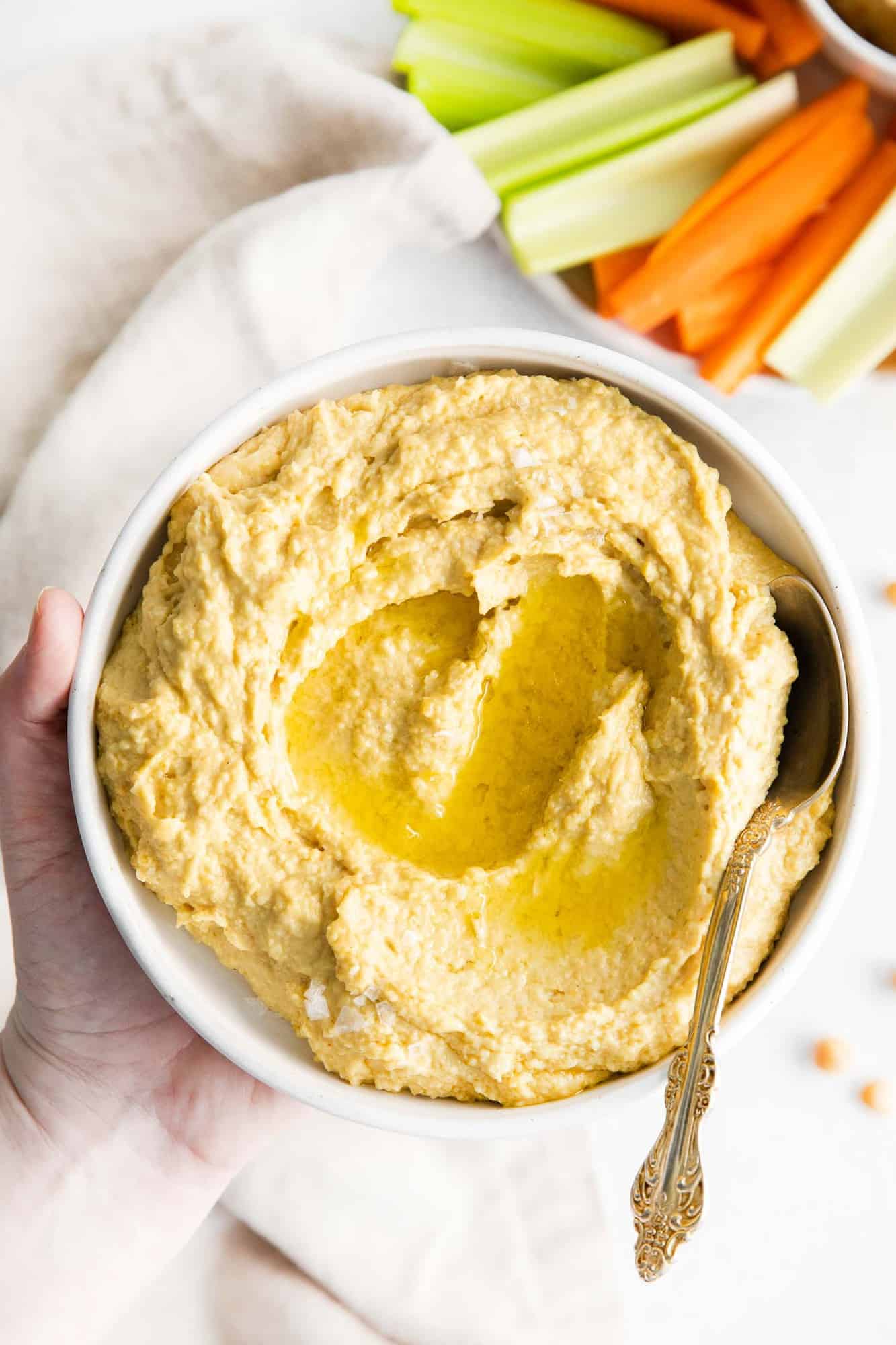 Bowl of hummus being held in a hand. Carrot sticks and celery sticks are also visible.