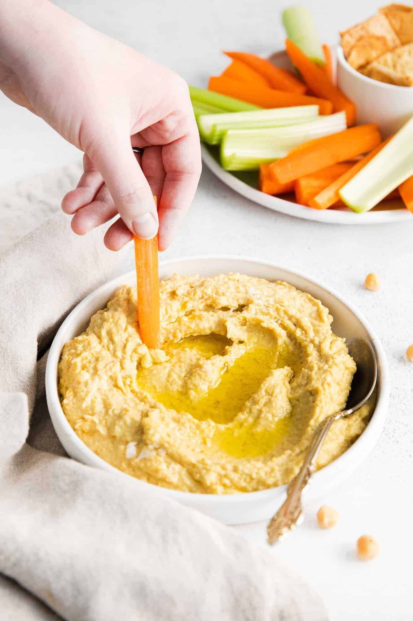 Hand dipping a carrot stick in a bowl of hummus.