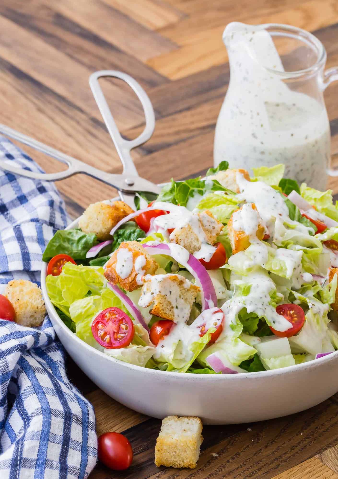 Tossed salad with tongs, small glass pitcher of creamy dressing in the background.