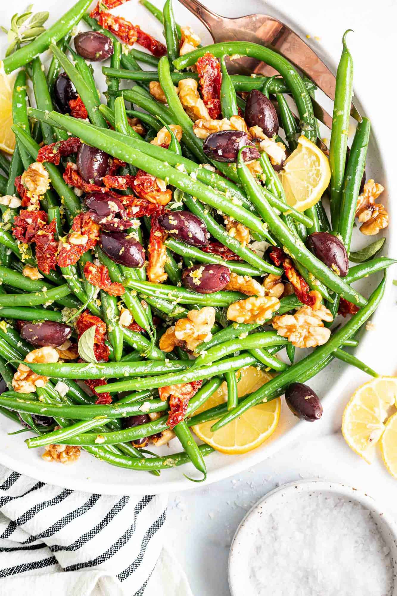 Green beans in a large white bowl with sun dried tomatoes, kalamata olives, and walnuts.