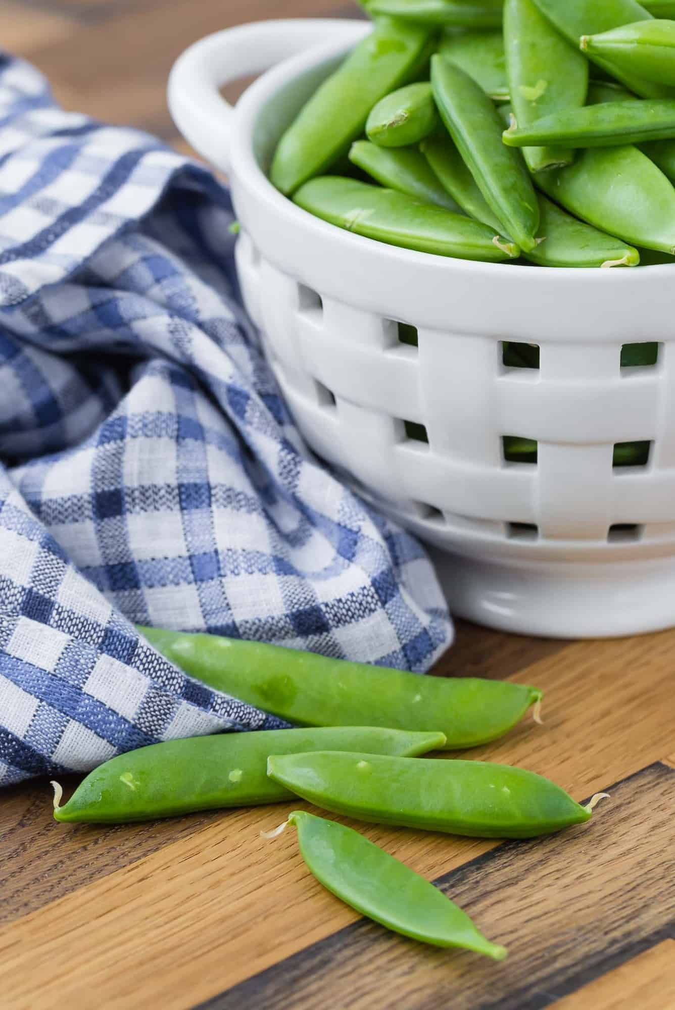 Sugar snap peas on a wooden surface in front of a small white strainer with more.