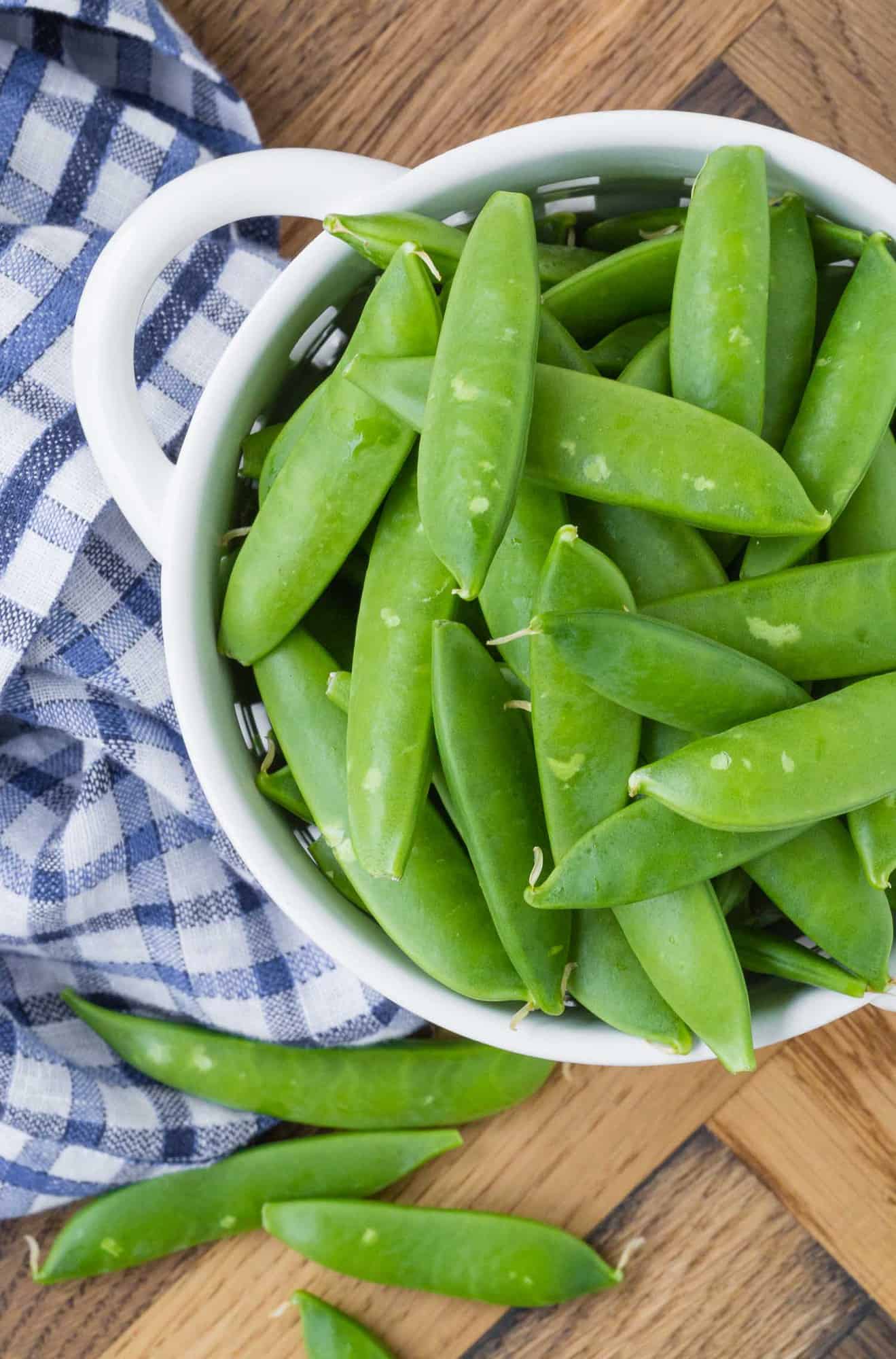 Overhead view of a small white strainer full of raw sugar snap peas.