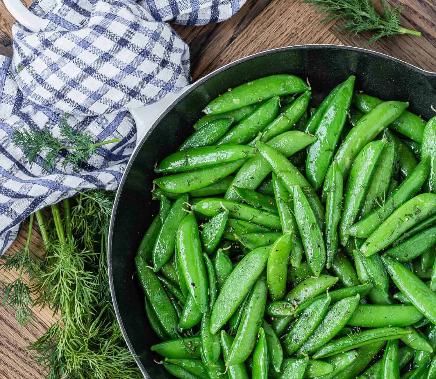 Cooked snap peas with dill in a pan on a wooden surface with a blue and white linen.