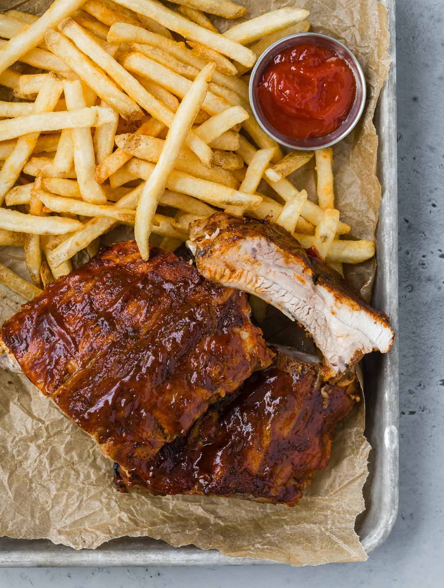 Ribs on a parchment paper lined tray with fries and a small metal cup of ketchup.