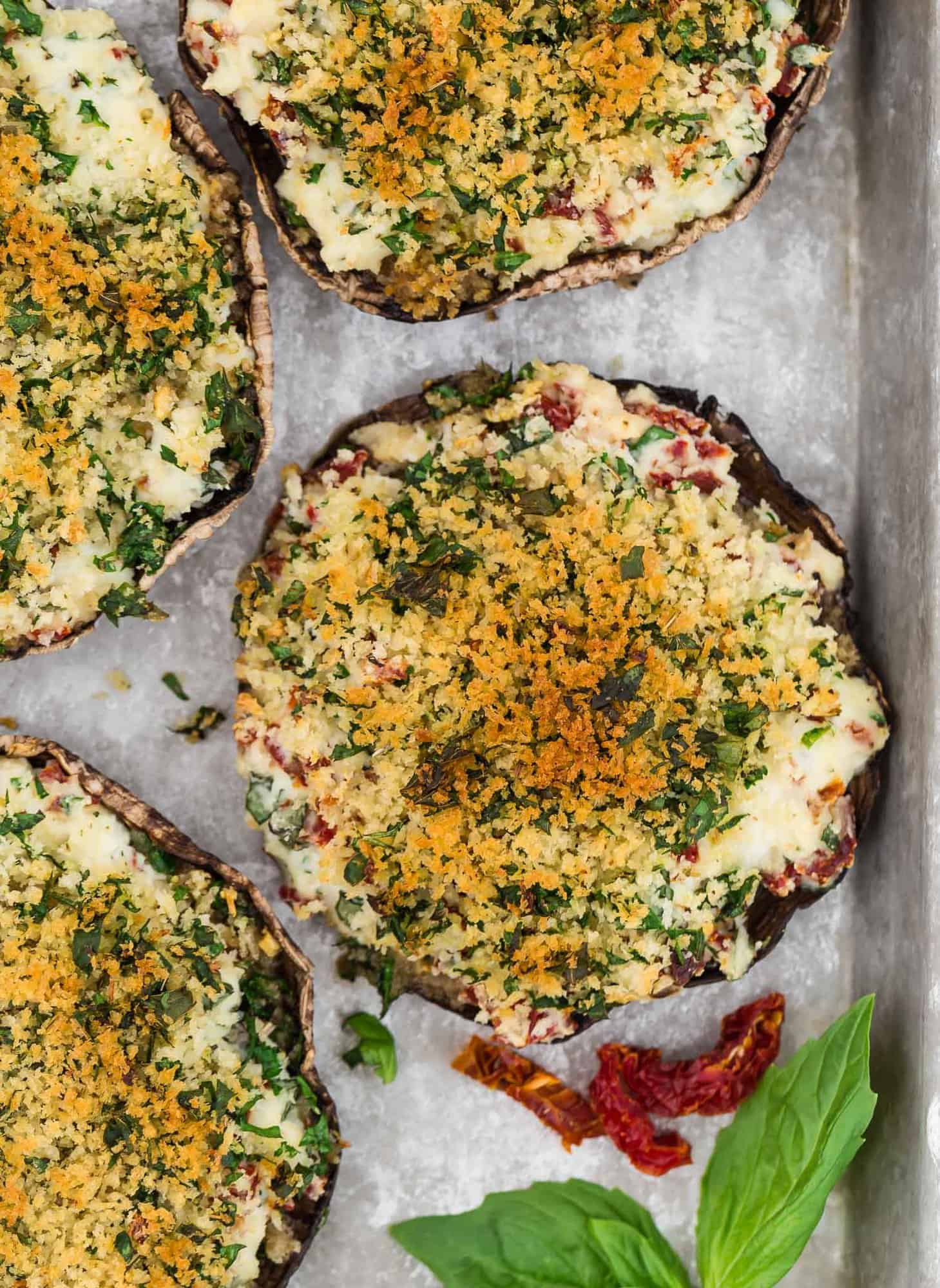 Overhead view of stuffed mushrooms on a baking sheet, garnished with sun dried tomatoes and fresh basil.