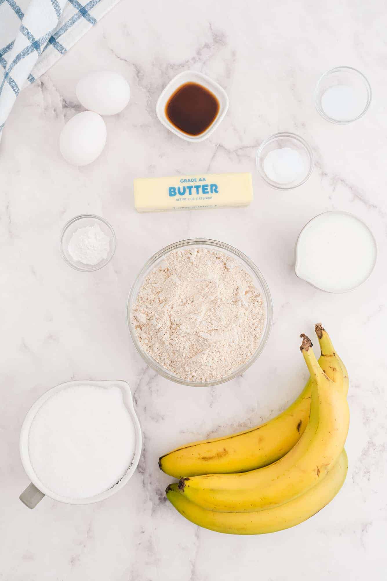 Ingredients for banana bread, including bananas, eggs, butter, flour, sugar, milk, vanilla extract, baking powder, and salt, arranged on a white marble surface.