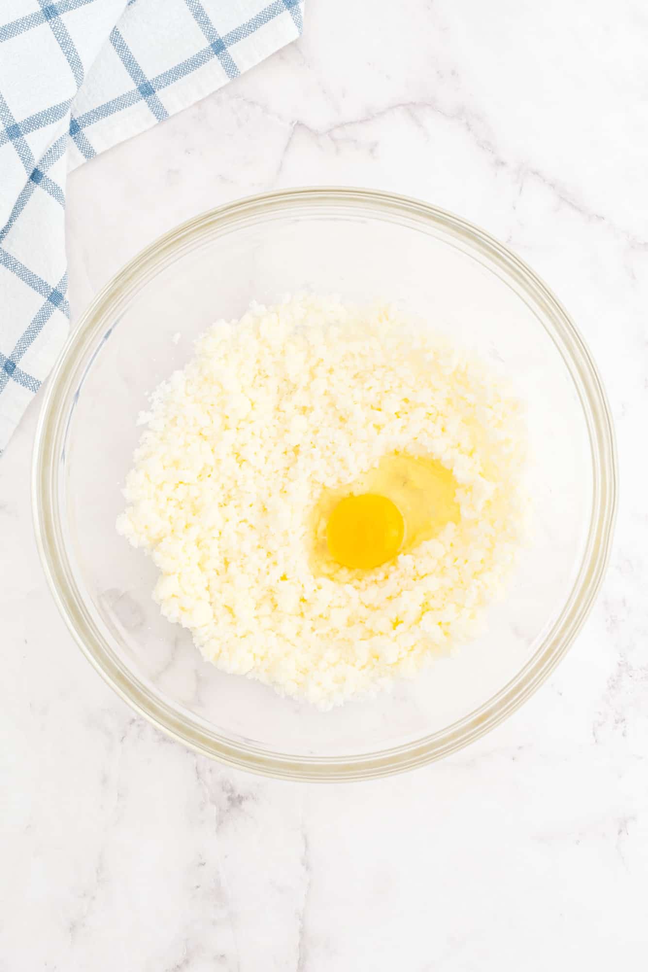 A glass bowl with butter and a raw egg sits on a marble countertop next to a blue and white checkered towel.