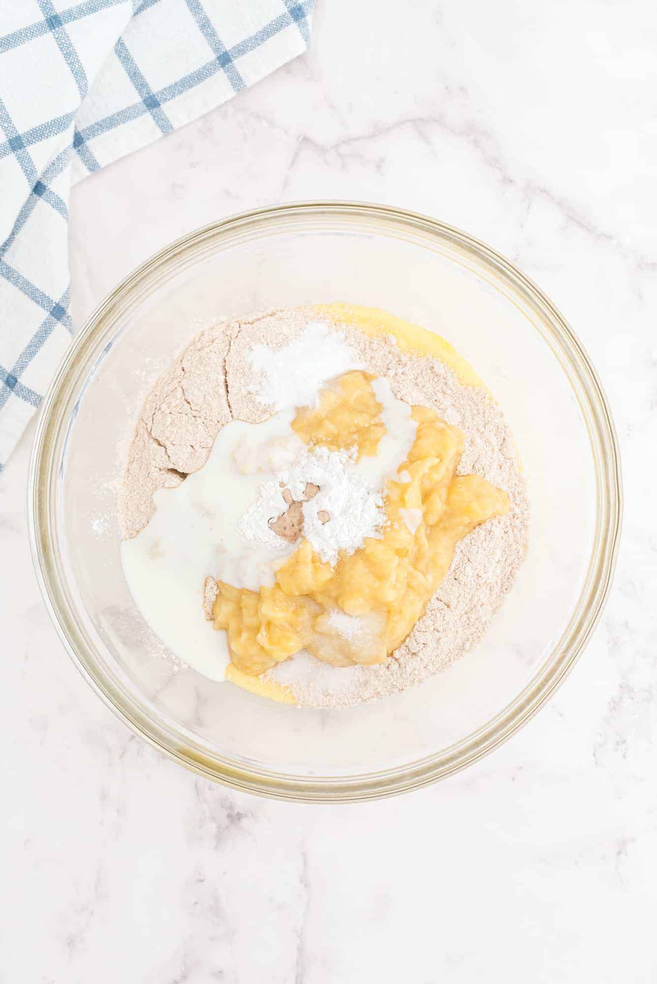 Clear glass bowl with flour, mashed banana, milk, and baking powder on a marble surface; a blue checkered towel is visible in the corner.