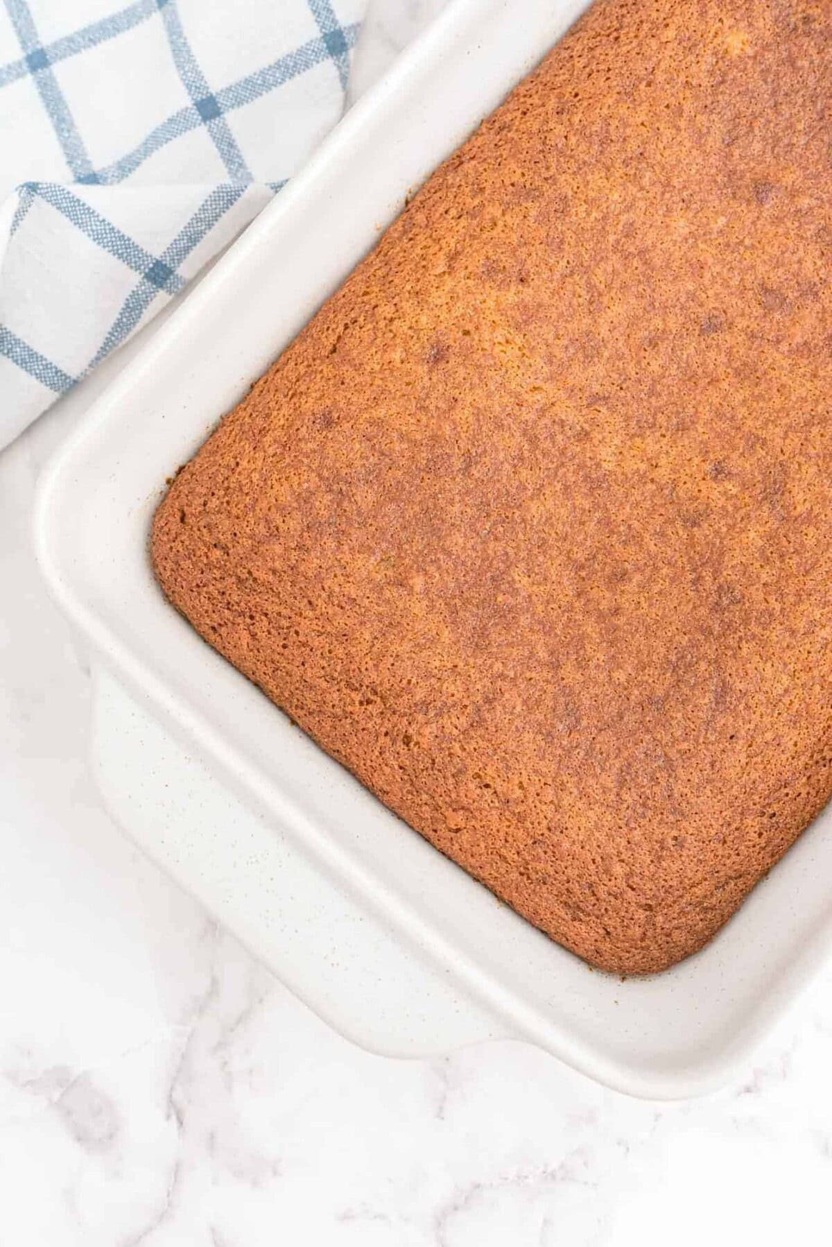 Rectangular, golden-brown cake in a white baking dish, with a blue and white checkered towel partially visible in the corner.
