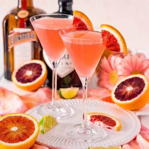 Two light pinkish red cocktails garnished with blood oranges, on a tray.